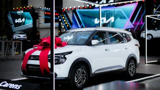 White car with a red bow on display at an automotive exhibition under bright lights.