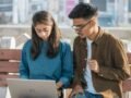 Two young adults working together on a laptop while sitting on a park bench outdoors.