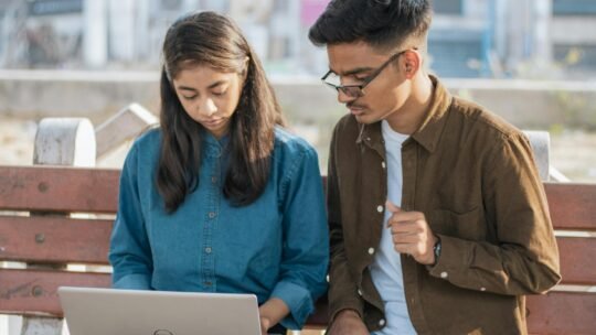 Two young adults working together on a laptop while sitting on a park bench outdoors.