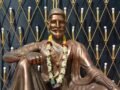 A bronze statue of Shivaji Maharaj adorned with a floral garland, set against a decorative backdrop.