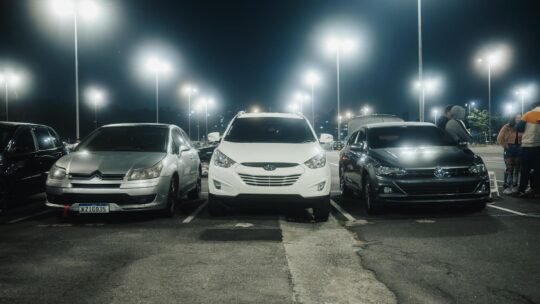 Night scene in a well-lit parking lot featuring various parked cars and street lamps.