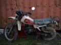A dirt-covered Honda 250R motorbike by a rusty container in Faenza, Italy.