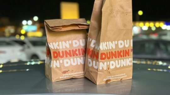 Two Dunkin' Donuts paper bags placed on a car hood with blurred night city lights in the background.