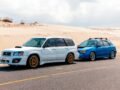 Two sporty Subaru cars parked on a desert road with sand dunes in the background.