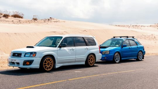 Two sporty Subaru cars parked on a desert road with sand dunes in the background.