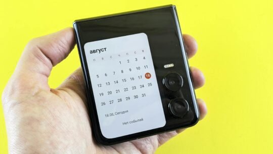 Close-up of a hand holding a flip smartphone with a calendar on screen against a vibrant yellow background.