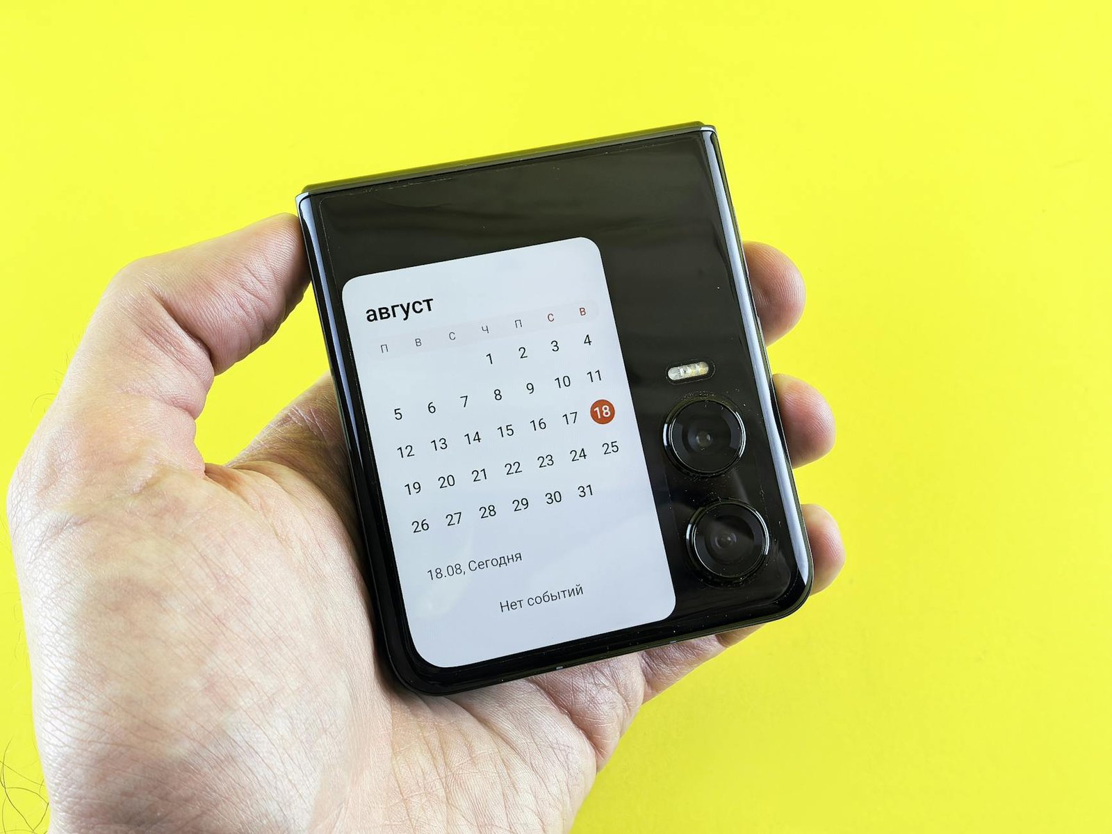 Close-up of a hand holding a flip smartphone with a calendar on screen against a vibrant yellow background.