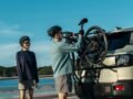 Two cyclists with helmets prepare their bikes on an SUV at a scenic coastal location.