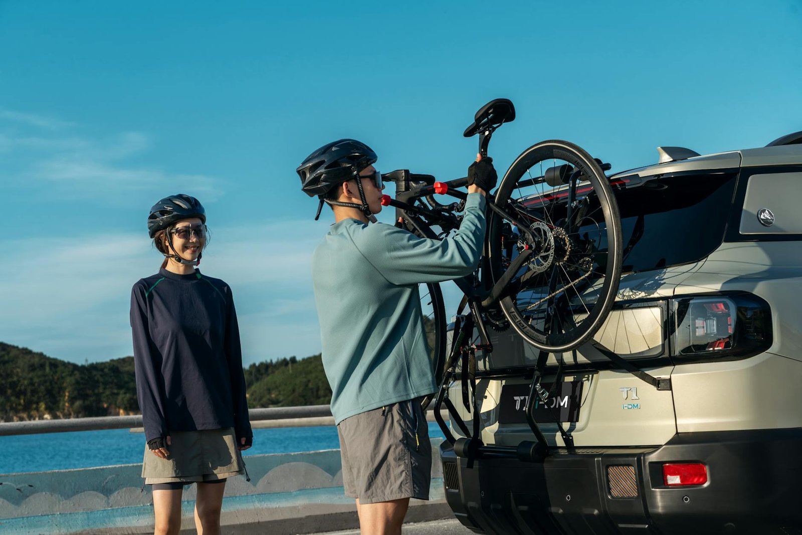 Two cyclists with helmets prepare their bikes on an SUV at a scenic coastal location.