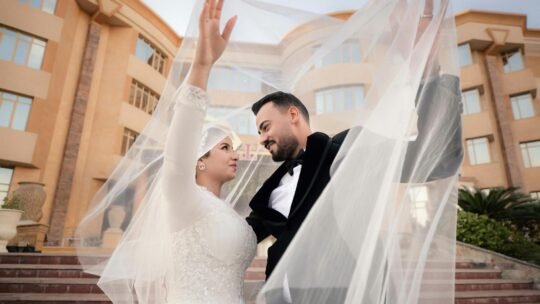 A couple celebrate their wedding outdoors, draped under a flowing veil with a grand building backdrop.