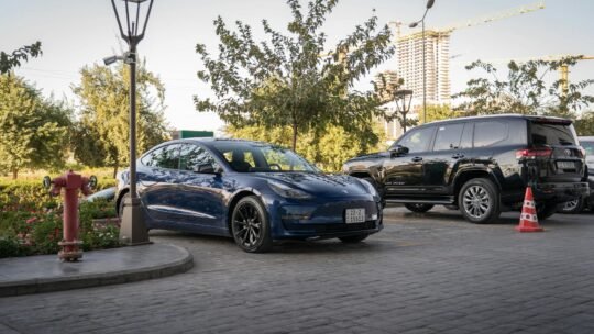 A sleek Tesla Model 3 electric car parked in a sunny urban parking lot with greenery and streetlights.