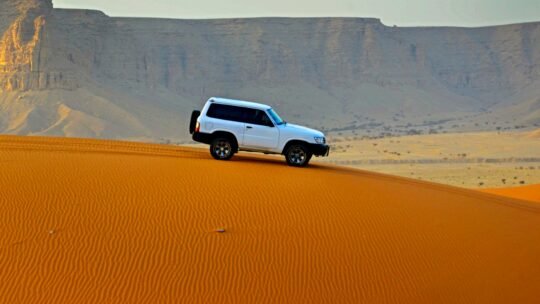 A white SUV navigates a steep desert sand dune at sunrise, with towering cliffs in the background.