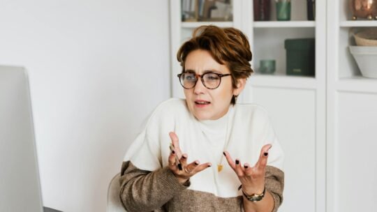 Expressive middle aged woman in casual outfit and eyeglasses gesticulating while having video conversation on computer in modern office