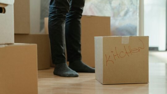 A person standing in a room surrounded by moving boxes labeled for the kitchen.
