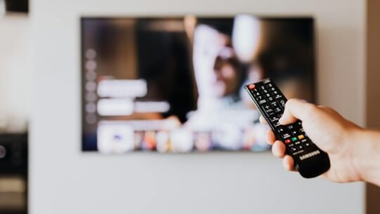 Person using a TV remote control with a blurred television screen in the background.
