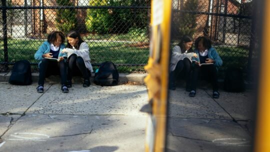 Full body happy multiracial girls classmates with textbooks sitting together on ground near school fence and chatting on sunny autumn weather
