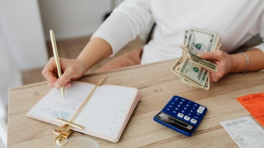 Adult holding cash and writing in planner while using a calculator at home.