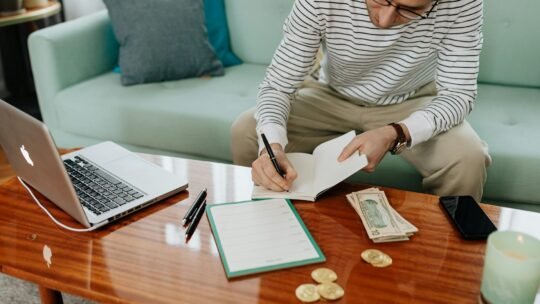 A man in a striped shirt sitting at a wooden table, writing in a notebook with money and a laptop nearby.