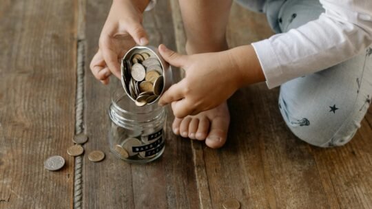 A child adds coins into a glass jar labeled for savings on a wooden floor.