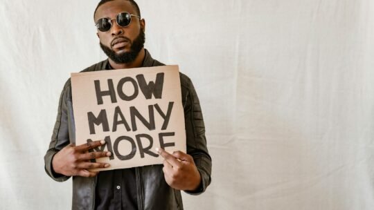 Man holding cardboard sign with 'How Many More' on a neutral background.