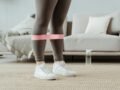 Close-up of a woman in sport leggings using a pink resistance band for leg exercises indoors.