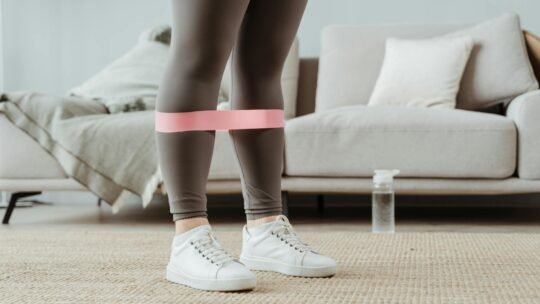 Close-up of a woman in sport leggings using a pink resistance band for leg exercises indoors.