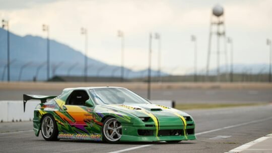 A colorful Mazda RX-7 parked on a racetrack with mountainous backdrop.
